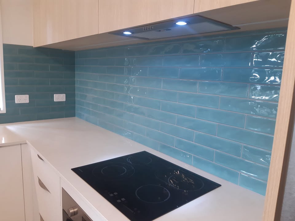 Glossy teal subway tile splashback above a white benchtop with induction cooktop and oak cabinetry, tiled by Angela Pullenger